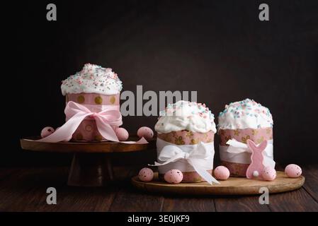 Festive Easter cakes with white icing and colorful sprinkles, wrapped in pink patterned paper and tied with pastel ribbons on dark wooden table Stock Photo