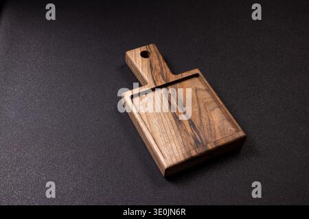 Walnut wooden cutting board with small handle and juice groove on a dark textured surface, minimalist rustic serving tray ready for food prep or prese Stock Photo