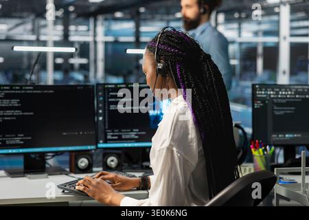 Data center operator using audio headset mic to provide user technical support. African American woman in server farm managing live support calls assisted by large language model chatbot Stock Photo