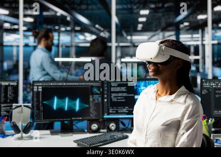 Smiling engineer using virtual reality, optimizing performance with AI assistant analysis. Cheerful data center female developer wearing VR headset, speed up tasks with LLM chatbot Stock Photo