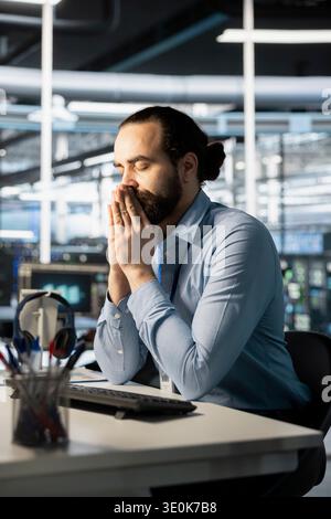 Exhausted server hub worker recollecting himself after repeated debugging attempts. Technician in data center sitting with eyes closed, managing stress after long hardware troubleshooting session Stock Photo