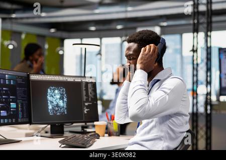 Technician optimizing performance using AI LLM chatbot, enjoying songs. African american IT expert putting on audio headset, using artificial intelligence assistant to automatize workflows Stock Photo
