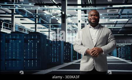 Smiling manager in data center doing checkup on hardware to avoid downtime. Happy african american supervisor overseeing server room infrastructure supercomputers, camera A Stock Photo