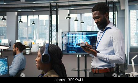 Technician next to office coworkers developing software applications, integrating artificial intelligence automation. African american man next to developers at work debugging applications, camera A Stock Photo