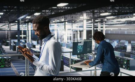 Software developers on data center industrial platform analyzing performance outputs on devices. Server room IT specialists collaborating on finetuning mainframes configurations, Stock Photo