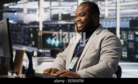 Joyous data center software developer delighted after solving all computer bugs. Smiling african american man in server hub pleased after finishing tasks on PC, overseeing server rigs, camera A Stock Photo