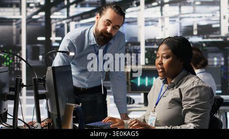 Server room manager providing feedback to employee managing AI training datasets. Team leader talking with technician in data center supervising system operations supporting neural networks, camera A Stock Photo