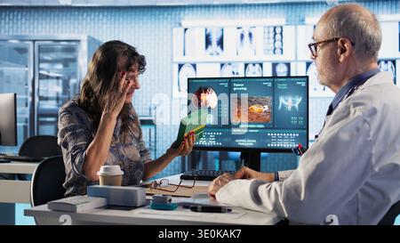 Old woman struggling with a severe migraine and asking for medicine, doctor putting up a recovery plan with strong painkillers treatment to relieve pain at checkup visit. Camera A. Stock Photo