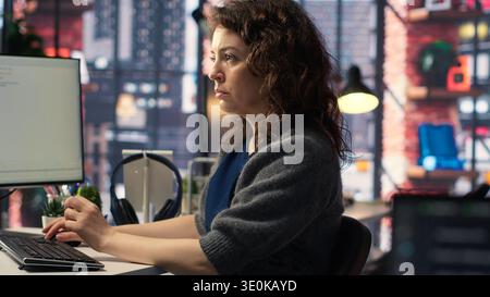 Young woman programming software code on her computer, using coding language for debugging tasks. Workspace with dual monitors helps with software engineering and web development. Camera B. Stock Photo