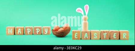 Minimalist Happy Easter banner featuring wooden blocks, a nest with speckled eggs, and a cute wooden peg doll with bunny ears on a vibrant green backg Stock Photo