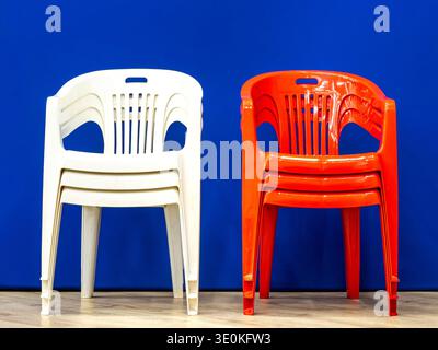 Front view of white and red plastic stacking chairs organized in two distinct piles on wooden floor against blue backdrop demonstrating color-coded furniture storage, selective focus Stock Photo