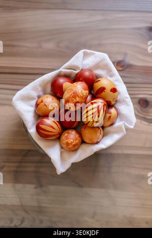 A basket lined with a white cloth holds several naturally dyed Easter eggs, featuring red and brown patterns created using onion peel. The eggs displa Stock Photo