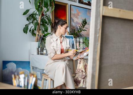 Focused woman artist drawing picture and enjoying art therapy, mixing colors on wooden oil palette. Stock Photo