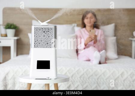 Modern air humidifier on table against woman using mobile phone in bedroom, closeup Stock Photo