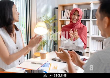 A diverse business team celebrates a successful project in a modern office. The Muslim woman wearing a red hijab smiles while clapping, showing teamwo Stock Photo