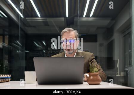 Middle aged Caucasian man wearing glasses sitting at desk working on laptop in modern office environment, looking at computer screen with focused expression Stock Photo