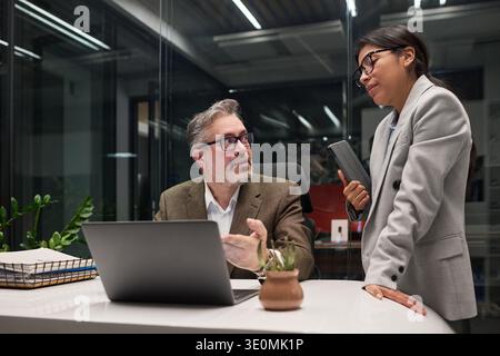 Middle aged Caucasian man sitting at desk discussing project with young adult Hispanic woman holding digital tablet in modern office setting, both engaged in conversation Stock Photo