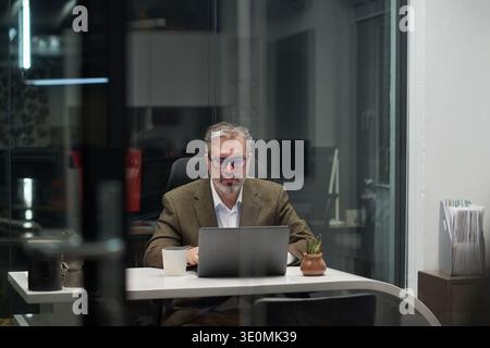 Middle aged Caucasian man sitting at desk working on laptop in modern office, wearing glasses and focused on screen, coffee mug and small plant placed on table nearby Stock Photo