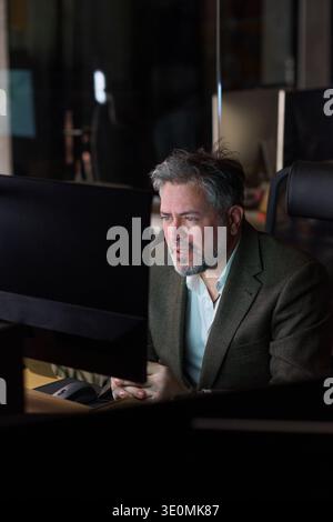 Middle aged Caucasian man sitting at desk working on computer monitor in modern office, facial hair visible, focused expression, hands clasped together, looking at screen Stock Photo
