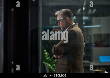 Middle aged Caucasian man standing with arms crossed in modern office environment, wearing glasses and looking down thoughtfully, indoor workspace visible in background Stock Photo