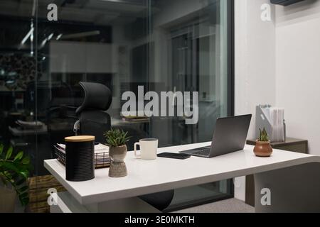 Modern office workspace featuring empty ergonomic chair positioned at desk with open laptop, coffee mug, smartphone, potted plants, stack of documents, glass wall background Stock Photo