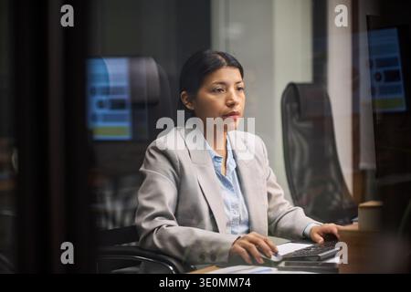 Young Hispanic woman working at computer in modern office, sitting at desk with focused expression, hands on keyboard, concentrating on screen Stock Photo