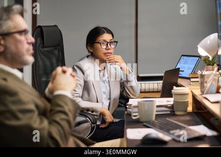 Young Latin woman wearing glasses sitting at desk in modern office, listening attentively during business meeting with middle aged Caucasian man, laptop and documents visible Stock Photo