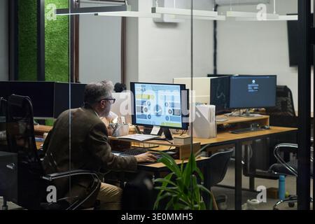 Middle aged Caucasian man working at desk using desktop computer with business analytics displayed on screen in modern office environment, sitting alone in workspace Stock Photo