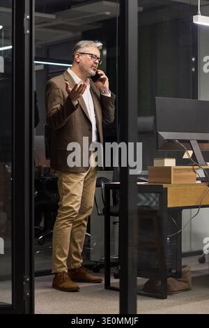 Middle aged Caucasian man standing in modern office talking on smartphone gesturing with hand near computer monitor appearing engaged in business conversation during workday Stock Photo
