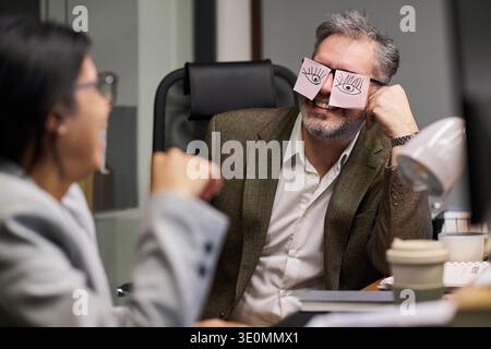 Middle aged Caucasian man sitting at office desk holding sticky notes with cartoon eyes over his own eyes, smiling at young Hispanic woman colleague laughing during conversation Stock Photo