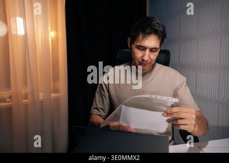Portrait of thoughtful man organizing essential paperwork in plastic sleeve, diligently working at home office desk with a laptop, managing his Stock Photo