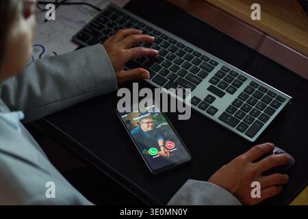 Man appearing on smartphone screen during video call, young woman sitting at desk using computer keyboard and mouse, hands visible interacting with devices Stock Photo