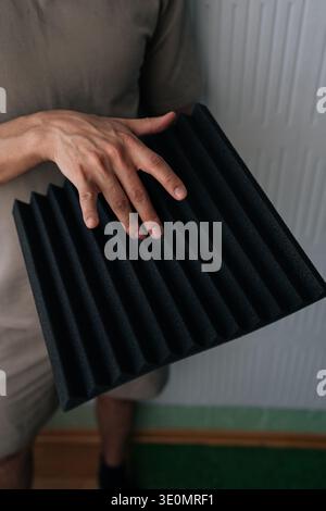 Close-up hand of handyman holding a black acoustic foam panel, exploring soundproofing materials for studio environment, noise reduction, and audio Stock Photo
