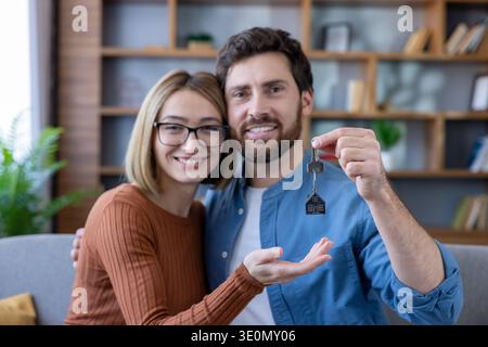 Smiling young couple embracing and celebrating new home ownership, proudly showing house keys with a charming home-shaped keychain, excited about their future together Stock Photo
