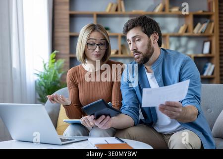 Young couple sitting on a sofa at home, managing household finances, calculating bills, reviewing papers, and experiencing stress about financial decisions and debt Stock Photo