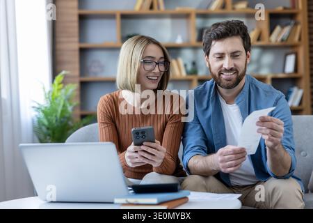Happy young couple budgeting household expenses, reviewing bills and payments using a laptop and mobile phone while sitting together on a sofa at home Stock Photo