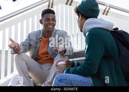 Male friends sitting on outdoor steps, talking and gesturing, holding coffee cup, carrying backpack Stock Photo