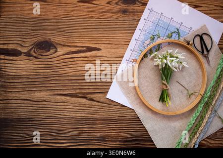 Embroidery hoop with linen fabric, cross stitch pattern and colorful embroidery threads on a wooden table with a small bouquet of snowdrop flowers. Co Stock Photo