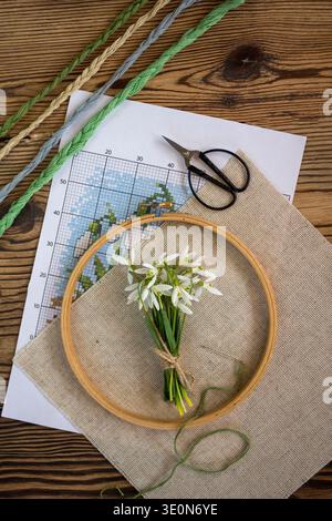 Embroidery hoop with linen fabric, cross stitch pattern and colorful embroidery threads on a wooden table with a small bouquet of snowdrop flowers. Stock Photo