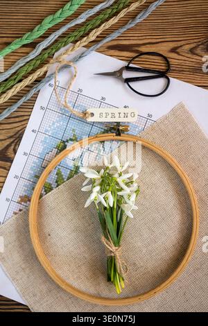 Embroidery hoop with linen fabric, cross stitch pattern and colorful embroidery threads on a wooden table with a small bouquet of snowdrop flowers. Stock Photo