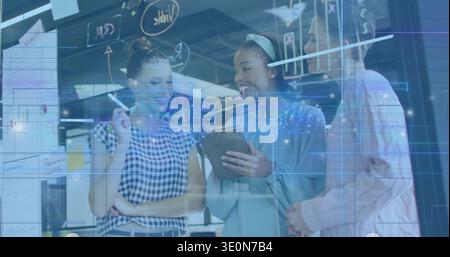 Diverse female coworkers collaborating at glass board in modern office, using tablet and marker Stock Photo