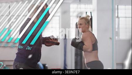 Punching female boxer in sports bra, boxing-gloved, training in lit gym, coach holding focus mitts Stock Photo