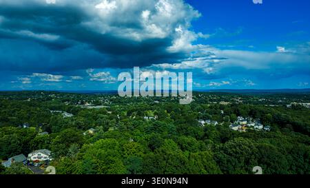 Aerial view of green town surrounded by forest under dramatic dark clouds and bright blue sky. Stock Photo