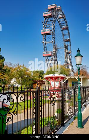 The Wiener Riesenrad Ferris wheel and a carousel stand in Prater amusement park under a bright blue sky, offering rides and views. Stock Photo