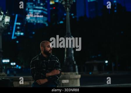 Bearded man sitting on a city bench at night looking thoughtful under street lamps Stock Photo
