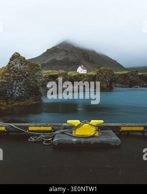 View of a solitary white house nestled near a rugged mountain partly hidden by mist, with dark blue water in the foreground, Arnarstapi, Snæfellsbær, Stock Photo