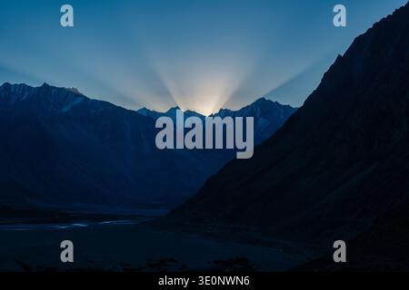 Bright sunbeams radiate from behind the dark, jagged silhouettes of the Karakoram mountains during sunrise in Diskit. Stock Photo