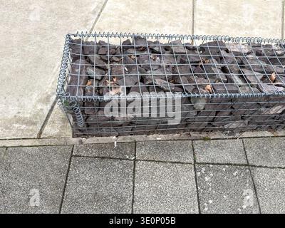 Close up view of a metal wire mesh gabion basket filled with dark stones resting against concrete paving slabs Stock Photo