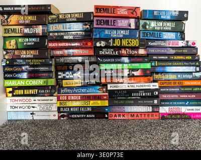 Large collection of thriller and crime paperback books stacked vertically on a carpeted surface with a white wall above Stock Photo