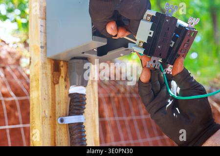 Worker attaches green wires to power panel while standing outdoors in construction area. Stock Photo